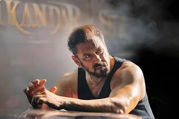 Bollywood superstar Salman Khan, wearing a black sleeveless shirt, leans forward with their hands clasped on a surface. The background is smoky, adding a dramatic effect. Partially visible text in the upper left corner seems to read "KANDAR."