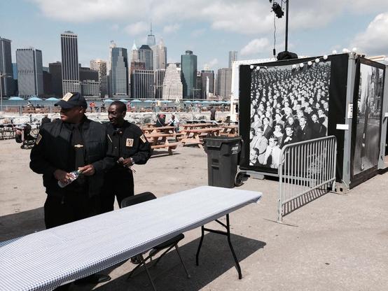 Photograph of two security guards looking off screen to the left while an image from Guy Debord's "Society of the Spectacle" book cover is displayed (the cover image is derived from a photograph taken in 1952 by the Life magazine photographer, J. R. Eyerman). The New York City skyline is in the background.