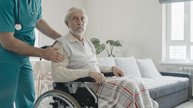 A healthcare professional in teal scrubs with a stethoscope gently supports an elderly man in a wheelchair. The elderly man, covered with a plaid blanket, looks off to the side with a mildly disdainful expression. They are in a well-lit, modern home setting with a sofa, houseplant, and large window in the background.