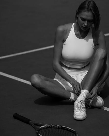 A woman sits on a tennis court, tying her shoelaces. She is wearing a white athletic outfit and white sneakers, with a tennis racket nearby. The image is in black and white.