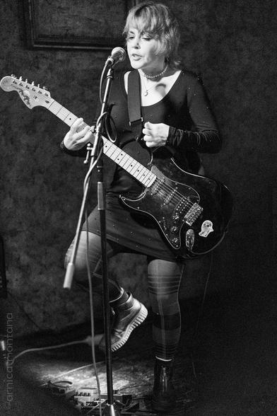 A musician performs on stage with an electric guitar. She is wearing a black dress and patterned leggings, and is singing into a microphone. The image is in black and white, capturing an intimate live music setting.
