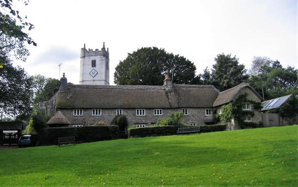 A row of thatched cottages with a village green laid out in front of them. Behind is the parish church with a rendered tower painted white rising up above the cottages.