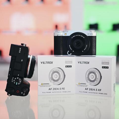 Display of two Viltrox AF 28mm f/4.5 prime lenses in their retail boxes, one for Sony E-mount (AF 28/4.5 FE) and one for Fujifilm X-mount (AF 28/4.5 XF). A Fujifilm X-M5 camera is positioned on top of the X-mount lens box, while a Sony A7C II camera with the Viltrox lens attached is standing vertically to the left. The background is blurred with vibrant colors, including orange and green panels.