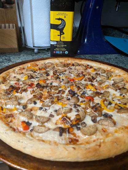A photograph of pizza topped with vegan cheese and sausage, button mushrooms, caramelized onions, and peppers. A bottle of Tunisian olive oil sits in the background.