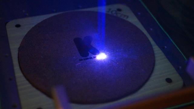 Lasergravur eines Korkuntersetzers mit einem blauen Laser. Graviert wird das Kindeko Logo.