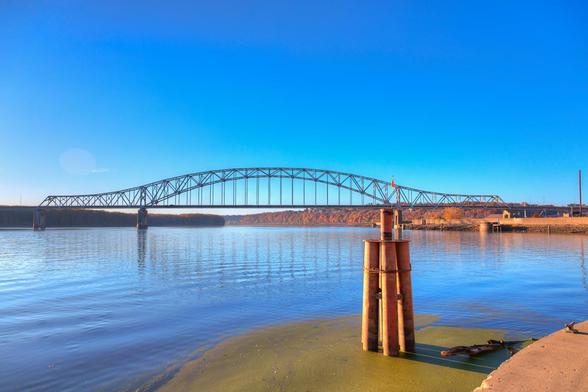 A steel bridge stretches across a large body of calm water under a clear blue sky.