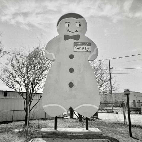 A black and white film photo. Smitty, a giant gingerbread man, hangs out at his roadside spot in Smithville, Texas. Saturday, March 22, 2025.