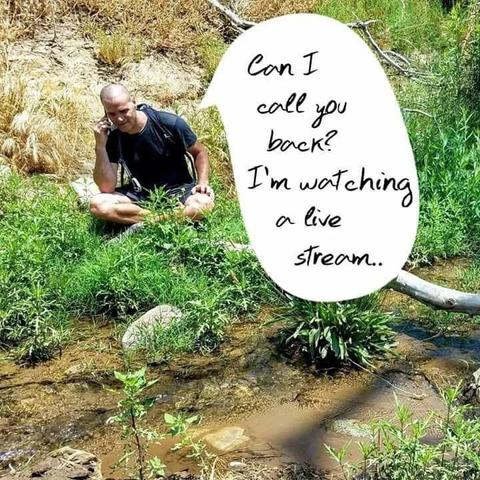 A man is sitting outdoors on the grass next to flowing water. He’s talking on a cell phone saying, “Can I call you back? I’m watching a live stream.”