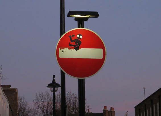 Ein Einfahrt-Verboten-Verkehrsschild, von einer Straßenleuchte beschienen. Es wurde von einem Künstler verändert, sodass nun eine schwarze, gespenstische Figur gezeigt wird, die hinter dem weißen Balken stehend diesen mit Hammer und Meißel beginnt abzutragen.