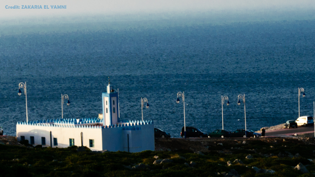 Blick über eine weiße Moschee mit blauen Verzierungen hinweg auf das dahinter liegende Meer, dessen Horizont im Dunst verschwimmt. Auf dem Meer ist weit und breit kein Boot oder Schiff zu sehen.
Oben links steht: 'Credit: ZAKARIA EL YAMNI'.