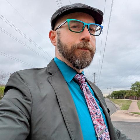 A photo of me, a 47 year old white man with a shaved head, a short cropped brown & grey beard, 

photographed from the chest up

In front of some live oak trees in an office park,

wearing

rectangular blue-green metallic acetate glasses, a navy & red paisley  necktie in a cape knot on a teal dress shirt under a charcoal waterproof sport coat and a matching flat cap.