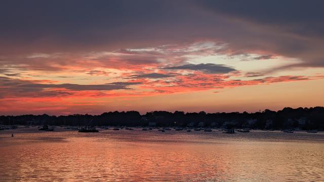 Historical Documentation in the form of a photograph of a rising sun over the ocean bay at the Historic Pioneer Village in Forest River Park, Salem, MA. The sky is filled with vibrant colors of orange, pink, and purple, with the sun rises in the background. The water is calm and reflects the colors of the sky, creating a mirror-like effect. In the foreground, there are several boats and yachts docked in the harbor. The harbor is surrounded by trees and buildings, and the horizon line is visible in the distance. The overall mood of the image is peaceful but electric.