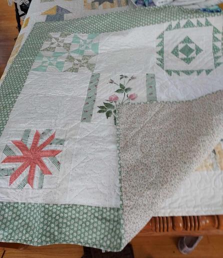 a quilt, it has a thick white and light green checkered border, in the middle is a white rectangle with a box in each corner in a unique design, lots of greens and light blues and some red and a flower in a box in the center, the corner of the quilt is turned up, showing the tight small floral design backing on khaki