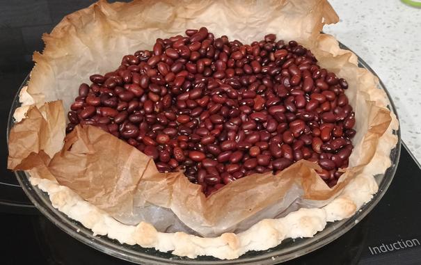 A pie shell in a glass pie plate is obscured by the parchment inside and filled with beans that hold it in place while pre-baking the pastry.