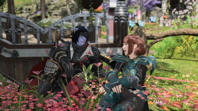 Lia and Esi sit on a bed of pink flowers chatting and laughing, under the shade of a tree.