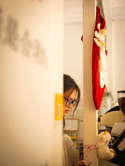 in a cozy indoor café, a girl wearing glasses is partially visible standing next to a white banner and serving coffee to her customer. near her, there's a red stocking decorated with a white reindeer pattern and a small plush toy. the overall atmosphere is warm and homely, with soft lighting contributing to a relaxed vibe.