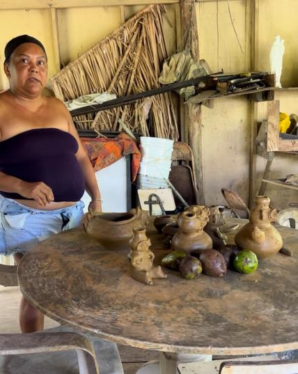 Potter Alice Cheverez, a brown skinned woman with her dark hair pulled back, wearing a tube top and jean shorts, stands behind a round wooden table that has pieces of her unfired pottery and ripening avocados sitting on it. There's a yellow wall and various items in the background. Additional info: Alice learned pottery from her mother, but her mother learned from a Taino cultural preservationist and archaeologist Daniel Silva, from Vieques.
