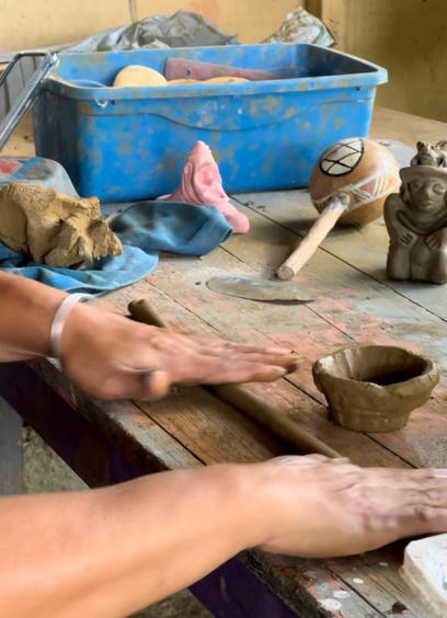 Alice's hands at her work table, rolling a coil of clay with her palms. A small cup of fresh clay, the beginning of a new pot, sits on the table in front of her. In the background there is a a small blue plastic bin, a lump of clay, a maraca and a clay figure she's already made.
