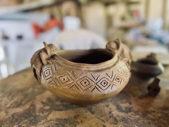 A finished piece that has not yet been fired. It is still the tan color of unfired clay. The piece is a deep bowl with geometric patterns carved along the front and back and small iguana figures on both sides. It is a good representative example of Taino pottery. The photograph is in portrait mode with everything but the pot blurred in the background.