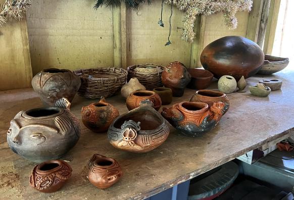 A table full of fired pottery. There are about a dozen pieces of different size and shape, mostly bowl forms with anthropomorphic figures attached to both sized, a common motif in Taino pottery. The unfired tan clay (from previous photo) has transformed to a brick red, with spots of black where the carbon from the smoke has entered the clay. 