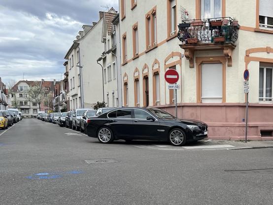 Une Berline BMW Serine 7 noire stationnée en diagonale sur le trottoir et le passage piéton au croisement de la rue Bastian et de la rue du Faucon, bloquant tout passage pour les piétons