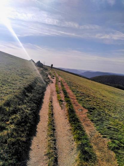 Das Bild zeigt einen Wanderweg, der sich durch eine hügelige Landschaft zieht. Der Weg ist unbefestigt und hat zwei parallele Spuren, die durch Grasflächen getrennt sind. Links und rechts des Weges erstrecken sich grüne Wiesen. Am Horizont sind sanfte Hügel oder Berge zu sehen, die mit Bäumen bewachsen sind.

Der Himmel ist teils bewölkt, mit sichtbaren Wolken, und die Sonne scheint von links oben herein, was einen strahlenden Lichtstrahl auf das Bild wirft. Auf dem Weg sind einige Wanderer zu erkennen, die in der Ferne gehen, was dem Bild eine friedliche und ruhige Atmosphäre verleiht.
