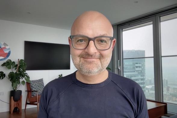 A selfie taken from a MacBook Pro, illustrating a tired face. In the background, a wall-mounted TV and a view of the neighbouring apartment block. 