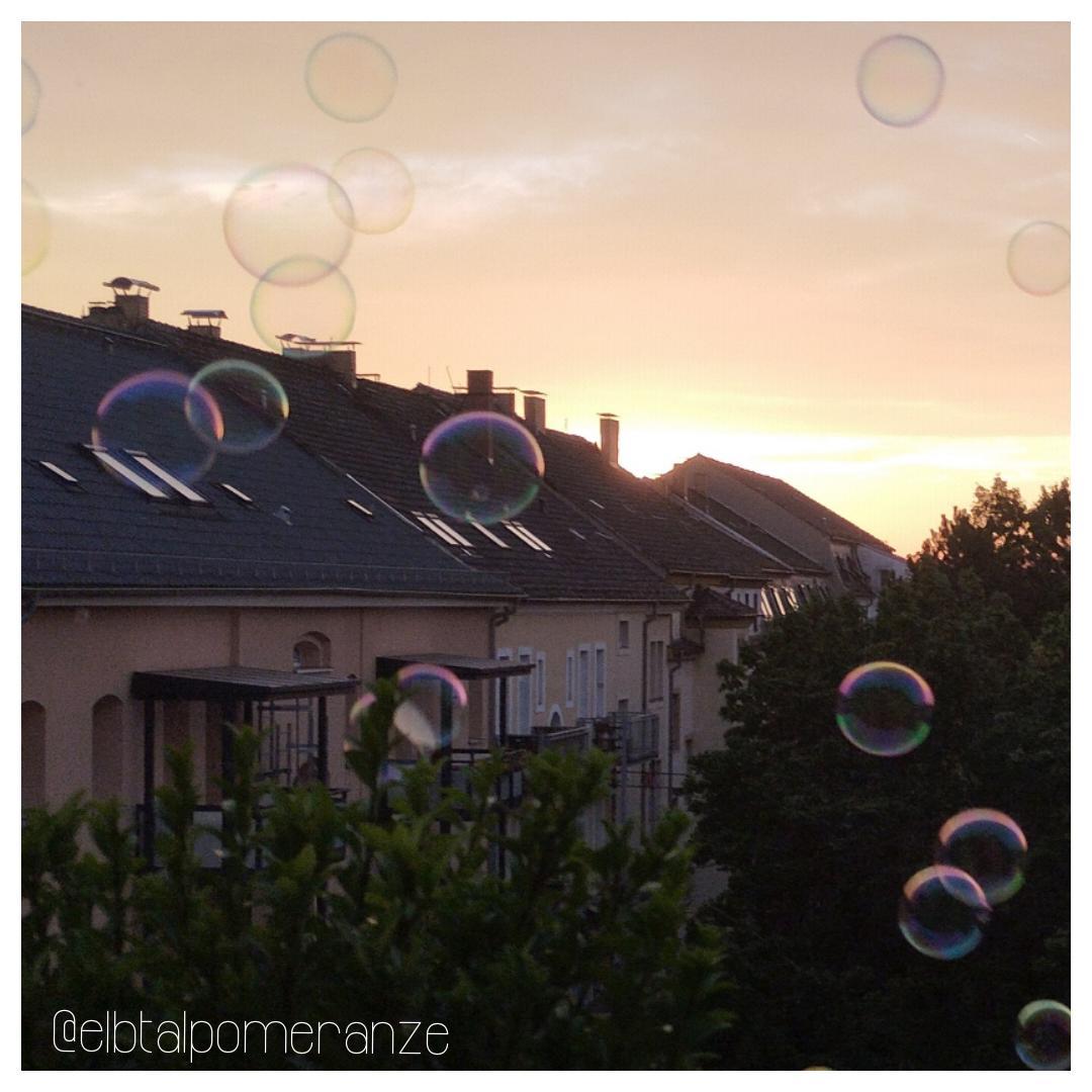 Blick von einer oberen Etage über einen von Mietshäusern (Altbauten) umrahmten Innenhof im letzten Abendsonnenlicht eines Sommertags. Zu sehen sind eine Reihe Häuser mit Balkonen, vor denen grüne Baumkronen die Sicht nach unten verdecken. Vor all dem schweben große, bunt schillernde Seifenblasen. Der Himmel changiert von oben nach unten von hellem Orange bis Gelb. Blick von einer oberen Etage über einen von Mietshäusern (Altbauten) umrahmten Innenhof im letzten Abendsonnenlicht eines Sommertags. Zu sehen sind eine Reihe Häuser mit Balkonen, vor denen grüne Baumkronen die Sicht nach unten verdecken. Vor all dem schweben große, bunt schillernde Seifenblasen. Der Himmel changiert von oben nach unten von hellem Orange bis Gelb.