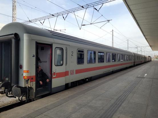 Former Deutsche Bahn carriage: white with a red lining and a "БДЖ" (BDZ) logo in place of "DB". The last door is open, with a staff worker sitting on the stairs waiting for arrival.