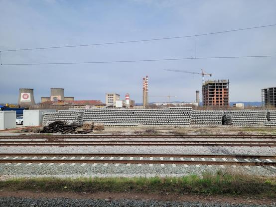 Stack of concrete railway sleepers (the beam between rails) after the last tracks. The Sofia thermal power plant is visible immediately behind, with 4 cooling towers, a handful of chimneys, and a couple of buildings in construction.