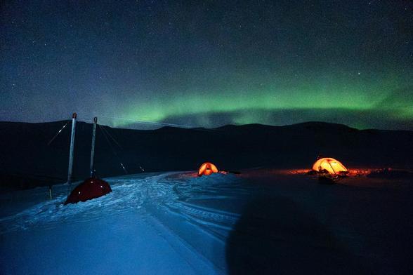 Photo of three red Hilleberg tents pitched on snow. It’s night, two tents illuminated, the one to the left is dark while some light is being thrown at it. There’s a bridge on the left, mountains in the background and bright green strip all across the dark sky. 