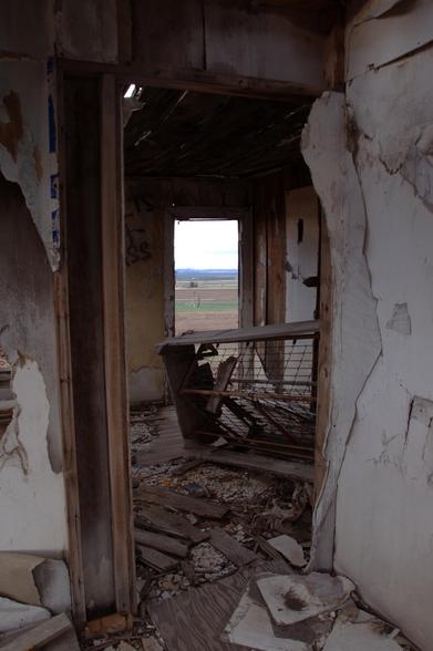 Looking through the doorway into the kitchen with deteriorating walls. Door to the outside can be seen with distant mountains and farmland