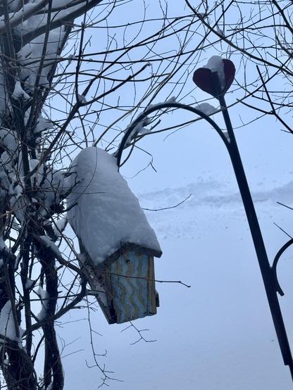 A close-up of a metal plant hanger with a heart at the top and a wooden bird feeder hanging from it with its roof piled with snow. 