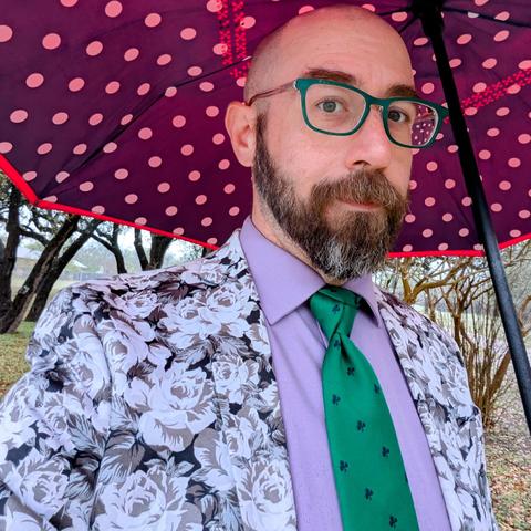 A photo of me, a 47 year old white man with a shaved head, a short cropped brown & grey beard, 

photographed from the chest up

Under a white on navy polkadot umbrella in front of trees in a park,

wearing

rectangular green acetate glasses, a green  necktie in a trinity knot on a lavender dress shirt under a greyscale rose print sport coat