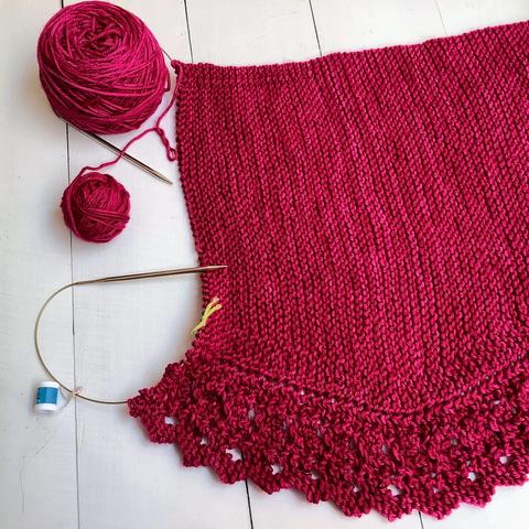 Angled view of a small ball as well as a cake of dark pink yarn, and a knitting project featuring garter stitch and lace on a circular needle. It's shown on a white plank background.
