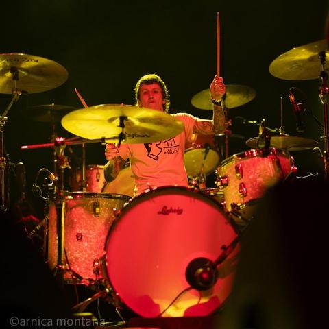 A drummer performs on stage, surrounded by drums and cymbals. The scene is lit with dramatic lighting, creating a vibrant atmosphere. The drummer is wearing a white shirt and appears focused on playing.