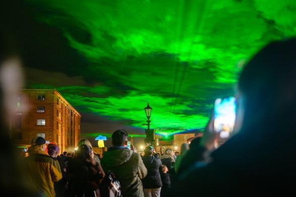 A crowd of people gathers at night in Liverpool's Royal Albert Dock to watch a light installation by Dan Acher called Borealis. The green light projection resembles the aurora borealis.