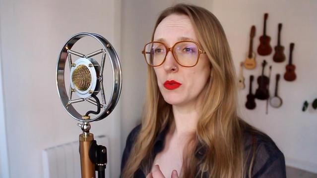Sage stands in front of a wall of ukuleles, singing in front of an old-style microphone. She is wearing bright red lipstick.