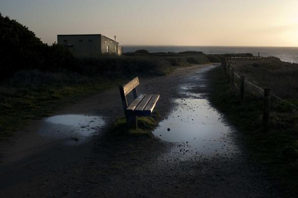 On est sur le sentier des douaniers, en Bretagne. Ce sentier côtier conduit du Mont-Saint-Michel à Saint-Nazaire. C’est la fin du jour. Un banc au milieu, face au soleil couchant, est éclairé par la droite. Des flaques devant et derrière ce banc reflètent la lumière qui baisse. Derrière le banc, plus loin en direction de l’horizon, un petit bâtiment blanc au toit plat reçoit lui aussi des derniers rayons de soleil. Seule sa face ouest est éclairée. Et pour horizon, l’Atlantique. 