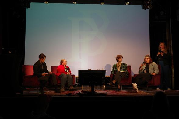 Photo: Four people are sitting on chairs on a stage, one person is holding a microphone. At the back right, a sign language interpreter.