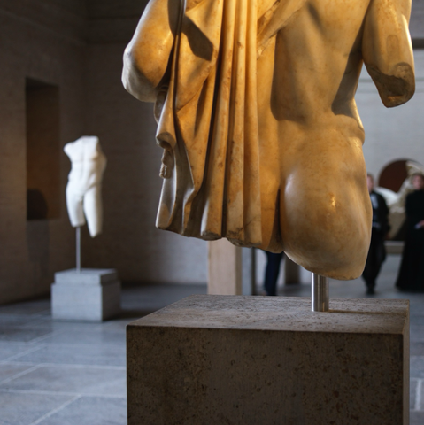 Partial, lower torso from behind of a classical marble statue. Focus is on the well-rounded bum that is partially concealed by the folds of a garment, the statue is carrying over their left shoulder. Unsharp in the background is a full torso statue some metres away. 