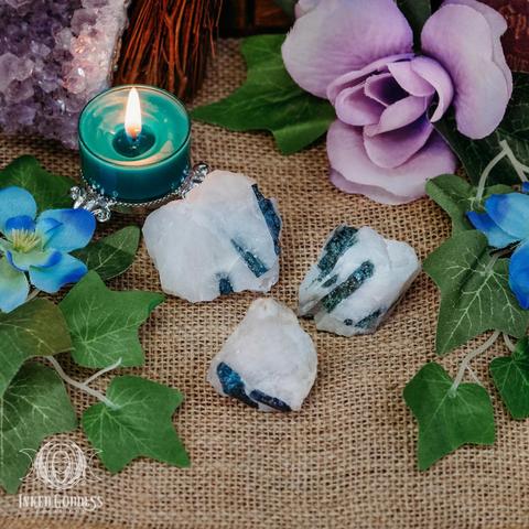 Three specimens of the "Indicolite on Quartz" product are arranged on a burlap-covered surface. The raw nuggets are mainly opaque or translucent white with bars of blue tourmaline (indicolite) visible on the surface. The one at the top left is larger while the other two are smaller and close in size. Presented by Inked Goddess Creations.