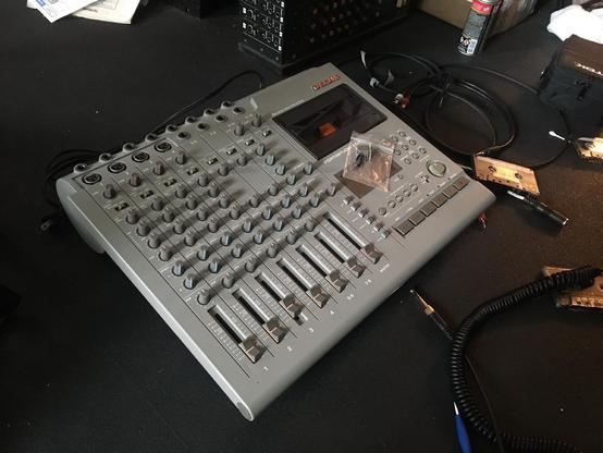 A Tascam 464 multitrack cassette recorder sits on a black rubber workshop floor, with cassettes and cables nearby.