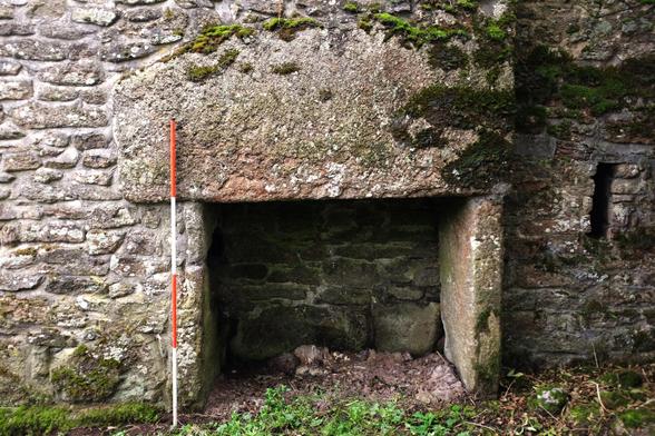 A medieval fireplace made of granite, with a very large single stone as a lintel. It is surrounded by smaller staones that make up the supports and the surrounding wall. To the right is a slit window.