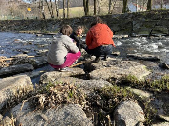 Foto einer Gruppe von drei Frauen die auf Steinen vor einem Flusslauf hockt und die in einer Konversation vertieft sind.