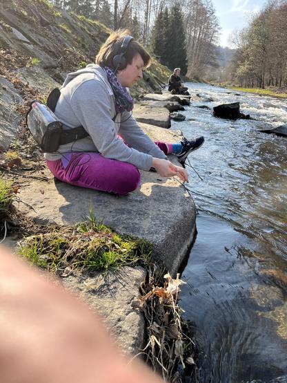 Im Vordergrund sitzt eine Frau mit Kopfhörern an einem Flussufer. Die Frau hat einen Recorder umhängen und ein Hydrophon (Unterwasser Mikrofon) in der Hand mit dem sie das Wasser aufnimmt. Im Hintergrund sitzt ein Mann und hört dem Wasser zu.