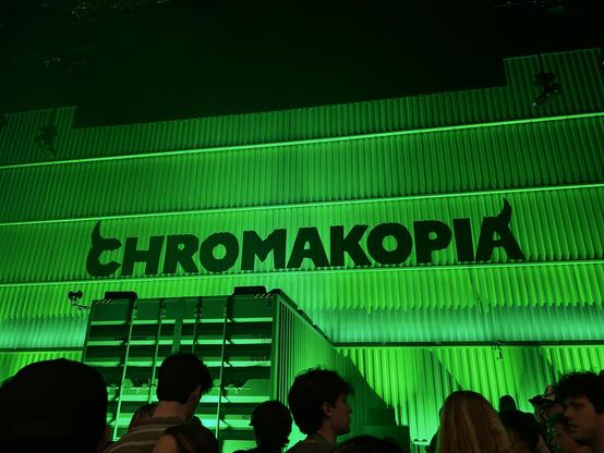 A crowd stands in front of a green-lit wall with the word “CHROMAKOPIA” displayed in bold black letters.