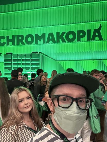 A man wearing a mask and glasses takes a selfie in a crowd at the CHROMAKOPIA event, with the green-lit sign visible in the background.