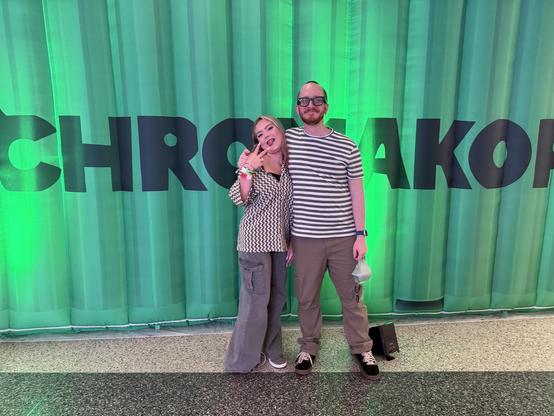 Two people pose in front of a large green CHROMAKOPIA backdrop; one is holding up a peace sign.