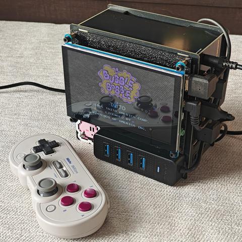 A black box shaped mini computer with various ports and black cables on the right side. The computer is on a beige rug on the floor. A small screen is attached to the front displaying the title screen of the game Bubble Bobble. Below the screen is a 4 port USB hub and a Kirby's Adventure Sticker. On the floor in front of the computer is an 8bit do wireless controller that resembles a Super Nintendo Controller.